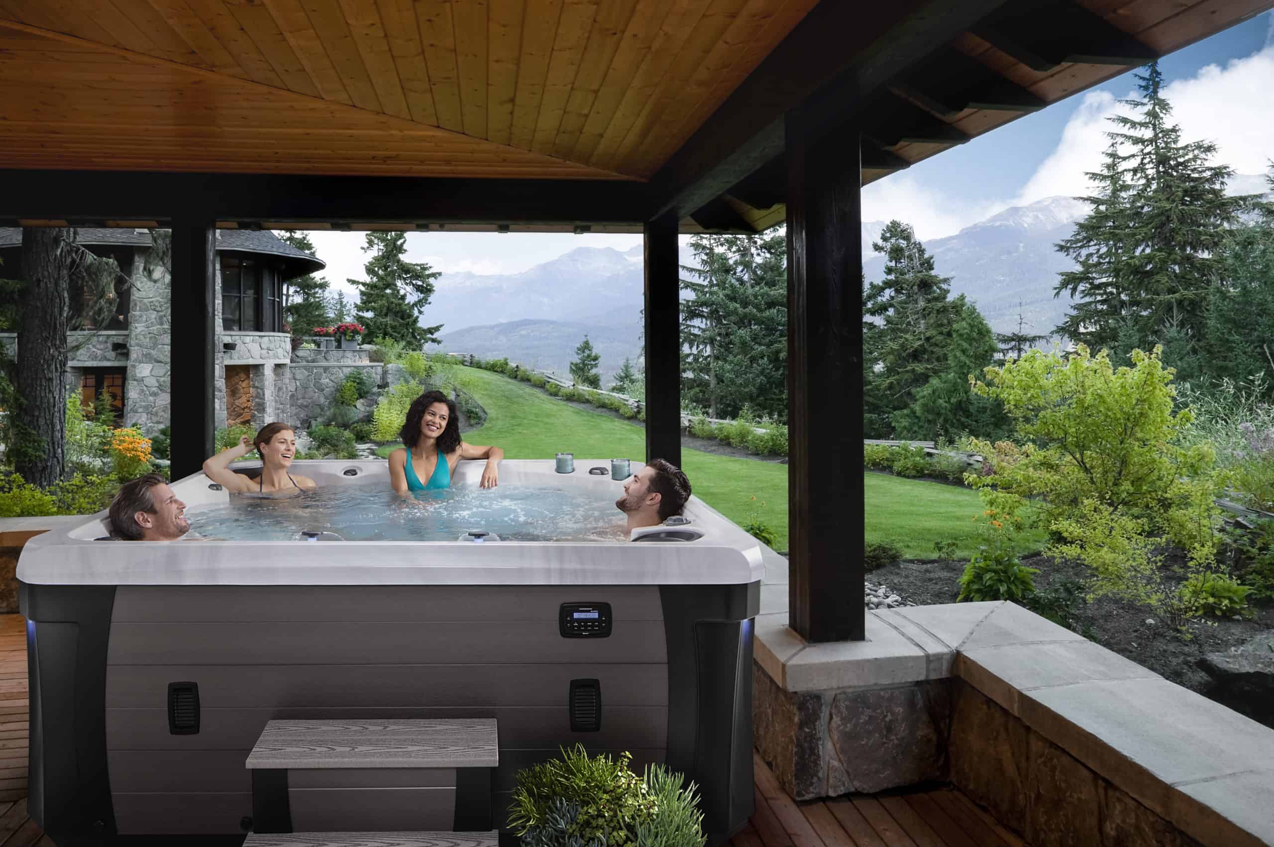 Two couples laugh and smile in a hot tub under a deck with beautiful mountains in the background as they discuss everything you need to know about buying and owning a hot tub in Idaho.