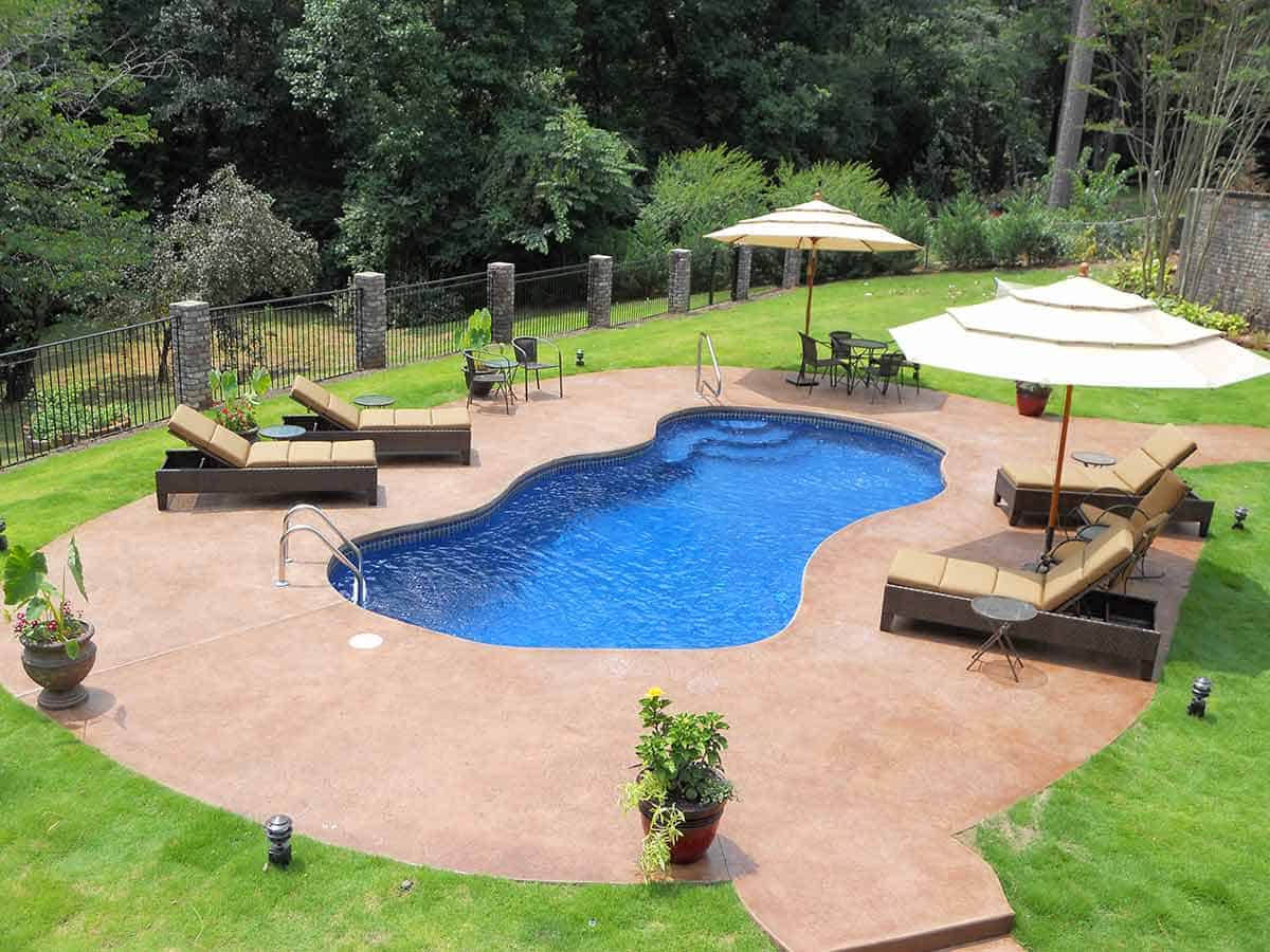 A beautiful fiberglass pool in Idaho sits surrounded by lounge chairs and umbrellas. Farther out are lush woods.