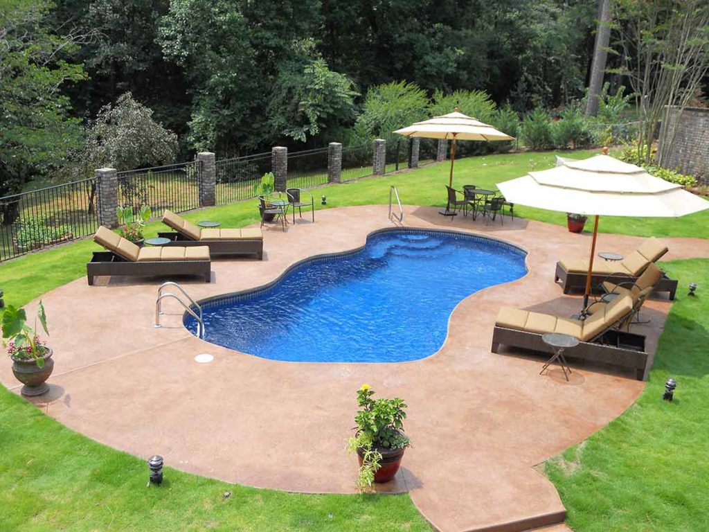 A beautiful example of fiberglass pools sit in the center of a poured concrete patio. Lounge chairs and umbrellas surround the fiberglass pool, and there are lush, green woods on the outer edge.