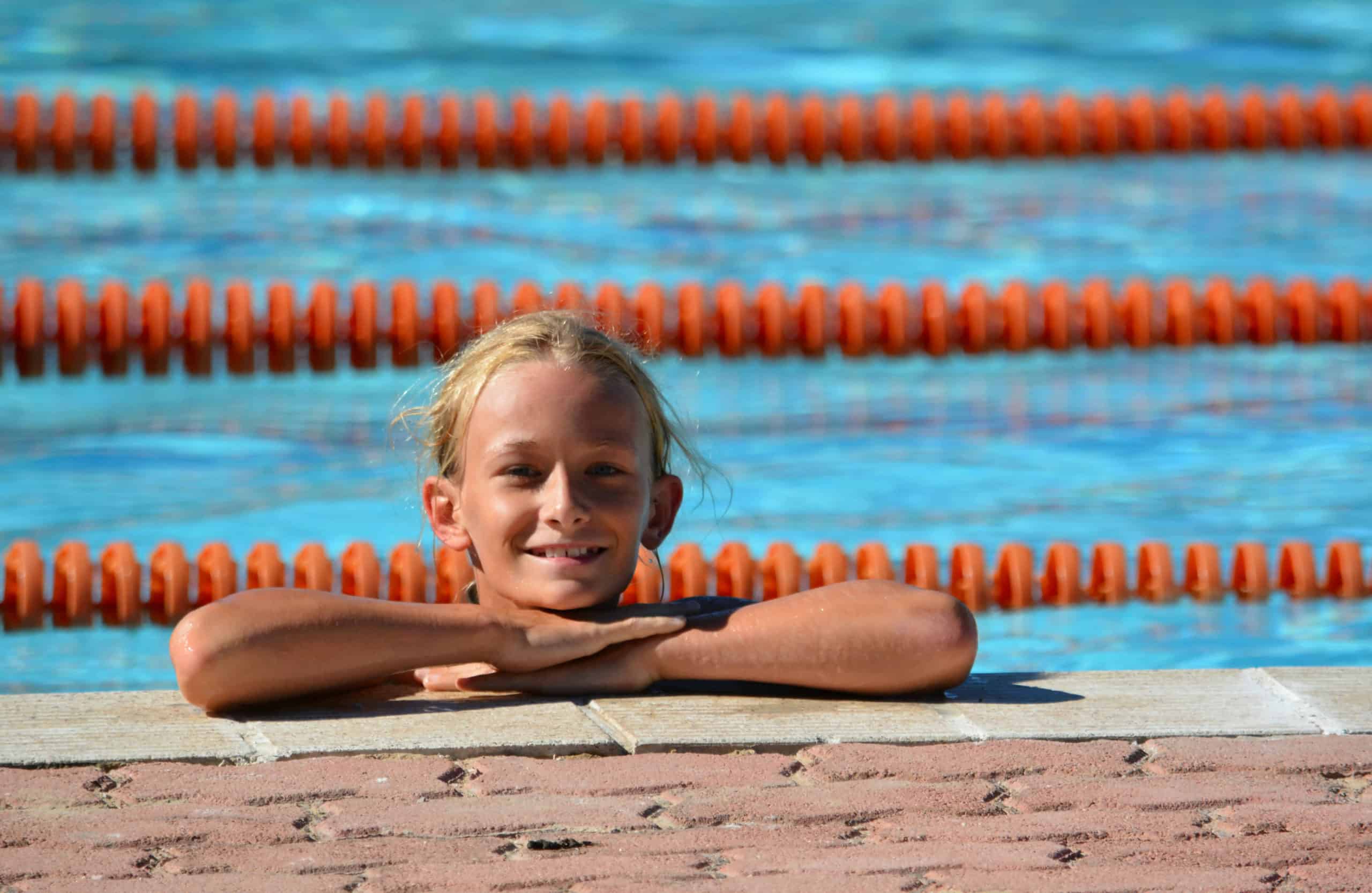 A girl floats in a public swimming pool that has commercial chemical delivery with her arms crossed on the edge. Orange swimming lane lines are in the background.