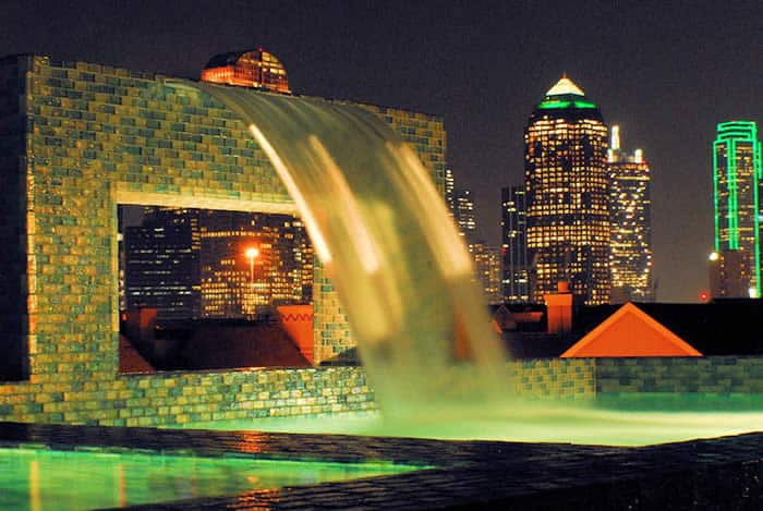 A luxury resot pool features a water cascade with a background of a nighttime city view.