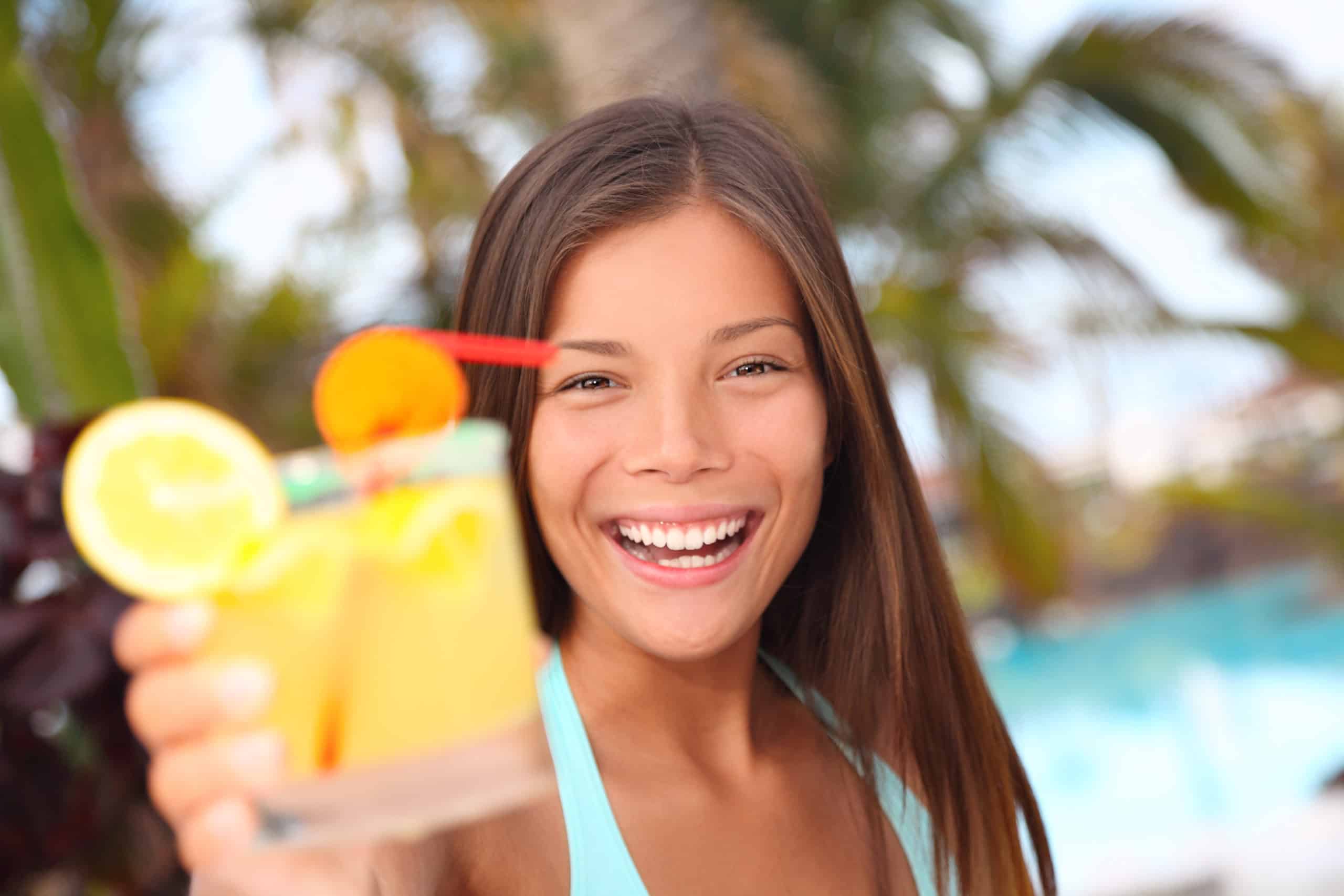 A woman with dark hair wearing a light blue swim suit holds a yellow, tropical drink as she is staying hydrated in her swimming pool.