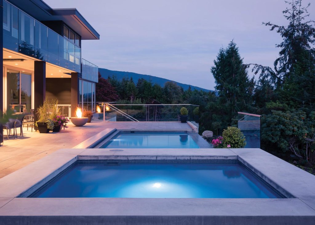 The homeowners of a lovely modern home with glass railings decided to instal a fiberglass pool. The pool is sleek and elegant, lit with gentle lighting as the sun sets over the mountain in the background.