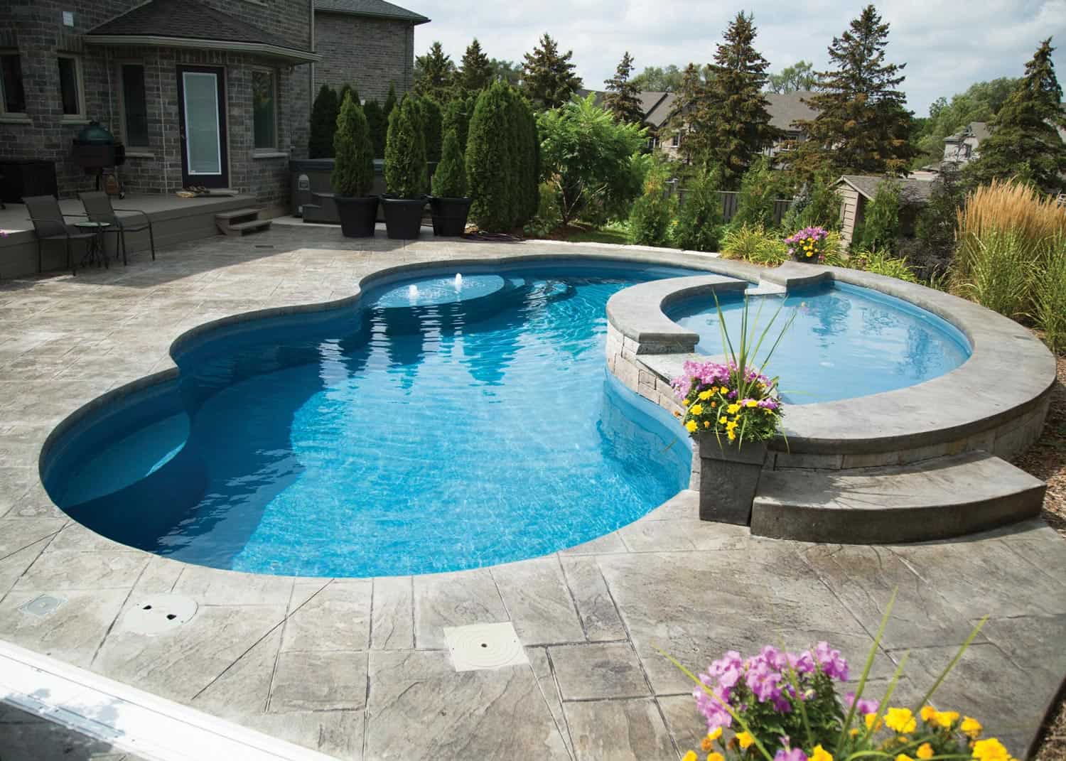 A beautiful fiberglass pool sits within a concrete pool deck as part of a well-done custom pool build. There is an attached spa with evergreen landscaping surrounding the pool area.