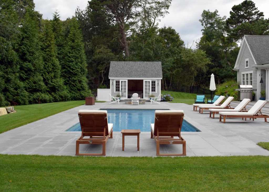 A beautiful, fiberglass pool sits nestled in a backyard full of evergreen trees with a small pool house across the pool. Closing your pool takes some planning.