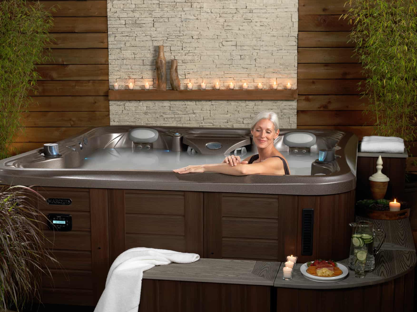Pictured is a woman in her at-home spa. She is sitting in a hot tub, her arm is along the edge of the tub and she is smiling at the camera. The hot tub appears to be in an enclosed patio and there is a stone backdrop with small candles. There is a bench in front of the tub with a towel, cucumber water, and a plate of food.