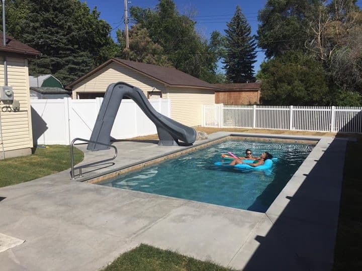 inground pool with two people floating on tubes in the water. There is a slide to the left with a handrail and stairs in the front left corner.