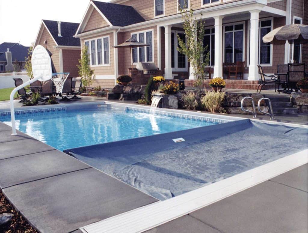 Automatic pool cover part way over an inground pool. The pool is located in a backyard and has a concrete path around it.