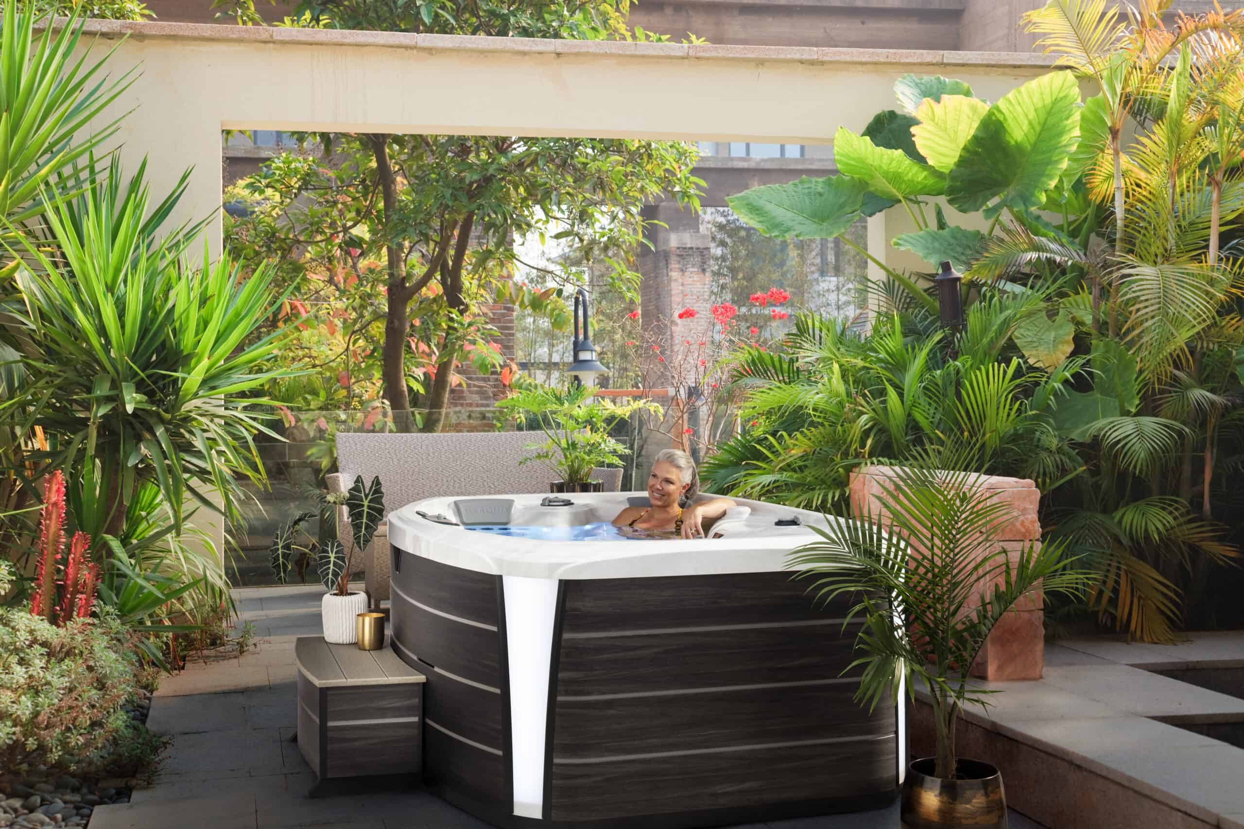 A woman is sitting in a hot tub practicing hot tub relaxation techniques by using the jets. The hot tub is surrounded by plants.