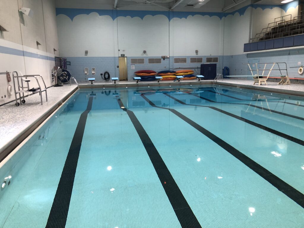 A commercial swimming pool is pictured in an indoor area. There are various pool items around the outside, and some stands to the upper right side.